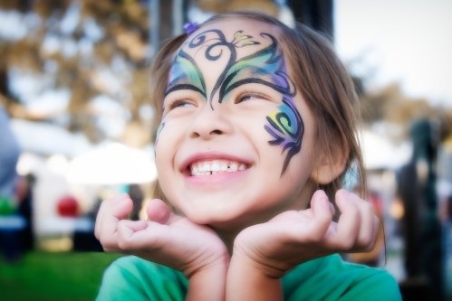 A smiling girl with colorful butterfly face paint, holding her hands under her chin, enjoying a festive atmosphere.