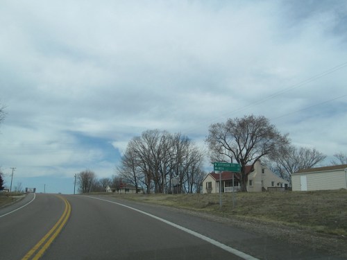 A winding road with a sign for a nearby town, flanked by bare trees and a few houses under a cloudy sky.
