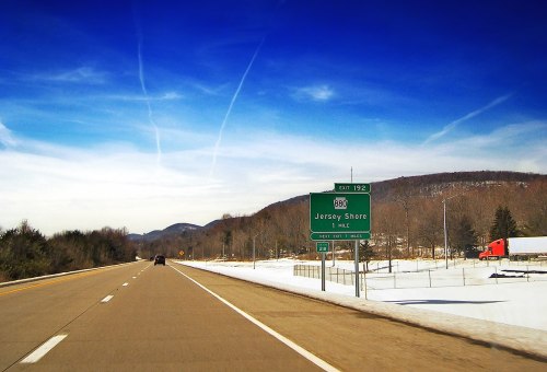 Highway scene with a sign for Jersey Shore, surrounded by snow-covered fields and mountains under a blue sky.