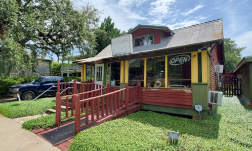 A colorful, quaint building with a sign that says "Open," surrounded by greenery and a blue truck parked nearby.