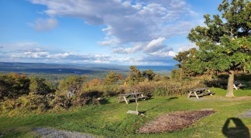 A scenic view of rolling hills and a blue sky with clouds, featuring picnic tables in a grassy area.