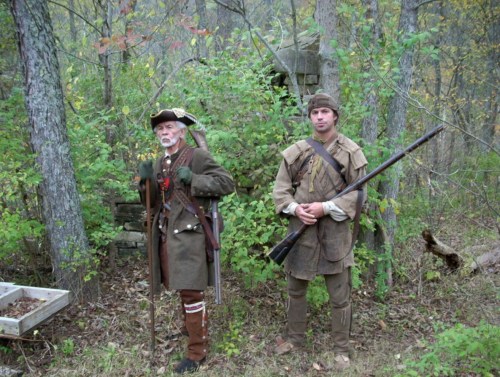 Two men in historical costumes stand in a wooded area, one dressed as a colonial soldier and the other as a frontiersman.