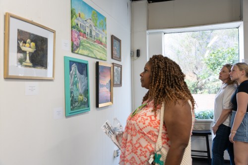 Three women admire various paintings displayed on a gallery wall, with natural light coming through a window.
