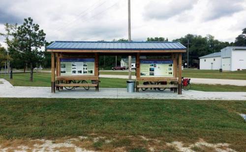 Two wooden shelters with informational panels and benches, set in a grassy area with a path and nearby buildings.