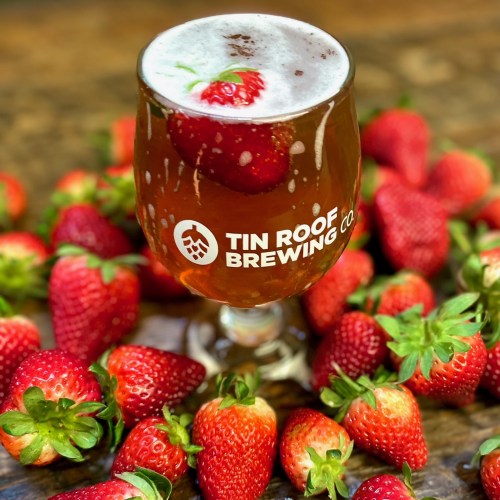 A glass of beer with a strawberry garnish, surrounded by fresh strawberries, featuring the Tin Roof Brewing Co. logo.