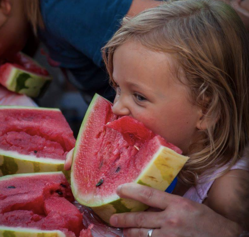 A young girl joyfully bites into a large slice of watermelon, surrounded by others enjoying the fruit.