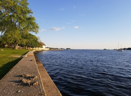 A serene lakeside view with a grassy area, trees, and boats on the water under a clear blue sky.