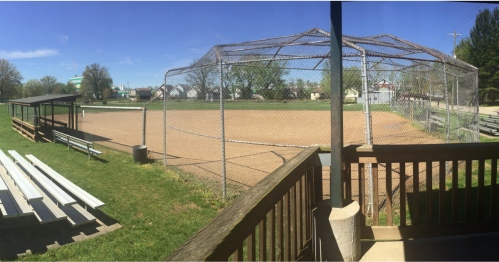 A panoramic view of a baseball field with a fenced diamond, bleachers, and a clear blue sky.
