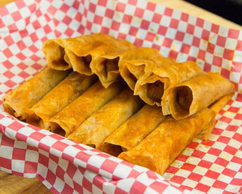 A basket lined with red and white checkered paper filled with golden-brown taquitos.