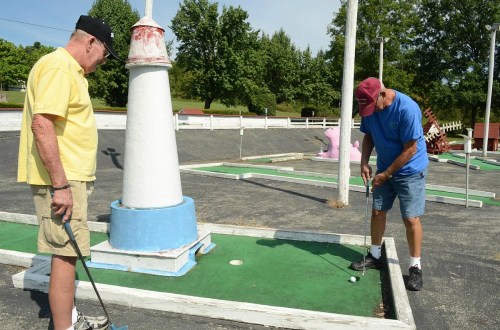 Two men playing mini-golf near a lighthouse structure on a sunny day, with trees in the background.