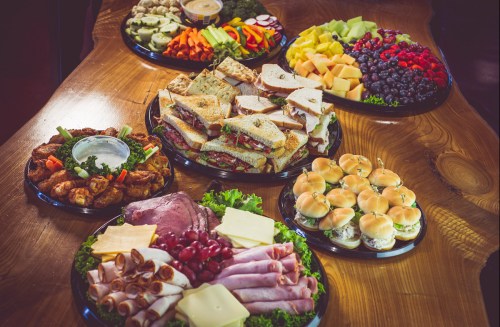 A variety of platters featuring sandwiches, meats, cheeses, fruits, and vegetables arranged on a wooden table.