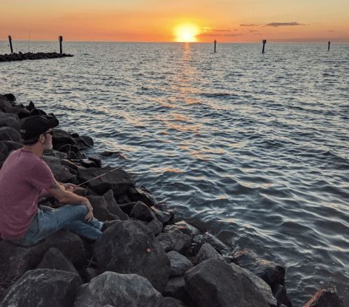 A person sits on rocks by the water, watching a sunset over the horizon.