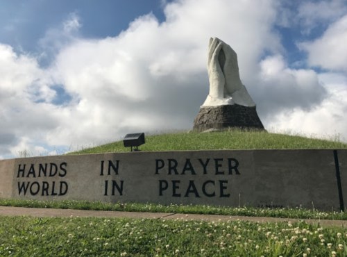 A large stone sculpture of hands in prayer, with the words "HANDS IN PRAYER, WORLD IN PEACE" on a grassy hill.