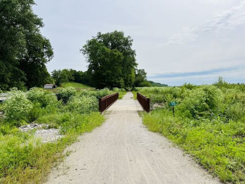 A gravel path leads to a metal bridge surrounded by lush greenery and trees under a cloudy sky.