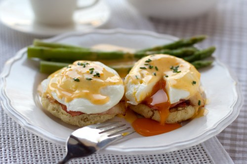 A plate with two eggs Benedict, topped with hollandaise sauce, alongside asparagus and a fork.