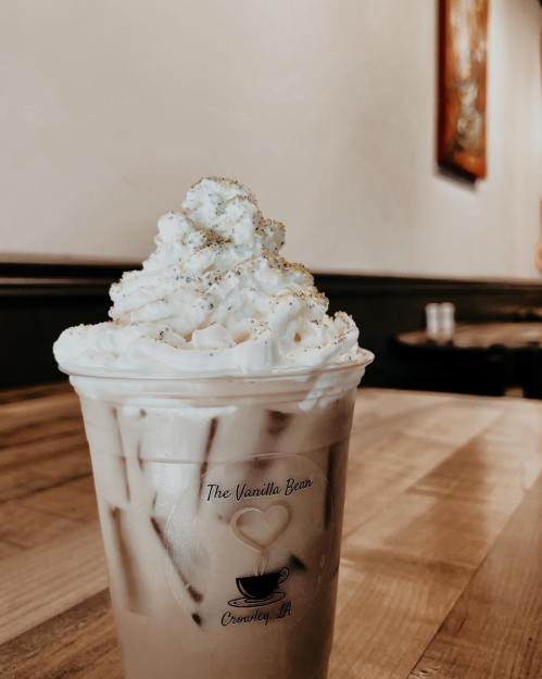 A close-up of an iced coffee topped with whipped cream and sprinkles, served in a clear cup on a wooden table.