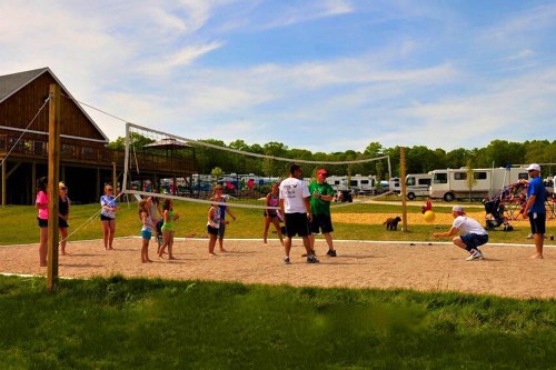 A group of people playing beach volleyball on a sandy court with a building and campers in the background.