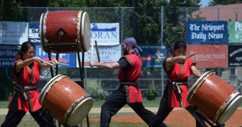 Three performers in red outfits play large drums outdoors, showcasing a dynamic and energetic drumming performance.