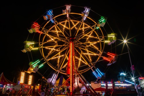A brightly lit Ferris wheel at night, surrounded by colorful carnival attractions and festive lights.