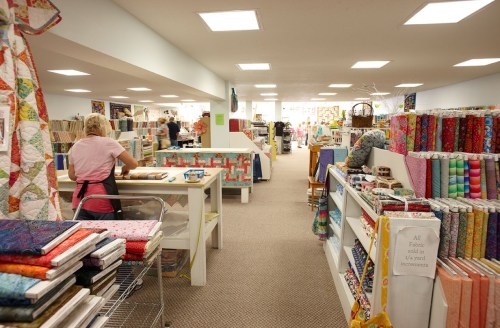 A bright fabric store with colorful fabrics, shelves, and customers browsing. A woman is working at a cutting table.