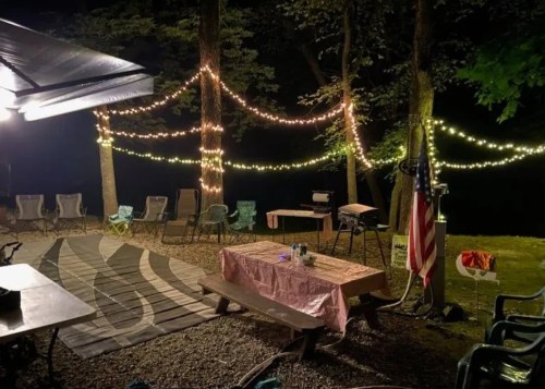 A cozy campsite at night, featuring string lights, chairs, a picnic table, and an American flag by the water.