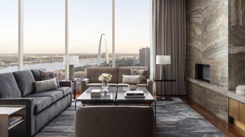 Modern living room with large windows showcasing the St. Louis Arch and river view, featuring stylish furniture and decor.