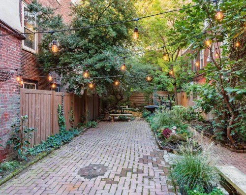 A charming brick-paved courtyard with string lights, surrounded by greenery and wooden fences.