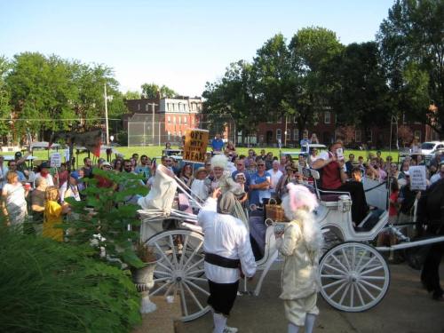 A crowd gathers around a horse-drawn carriage, holding signs and protesting, with trees and buildings in the background.