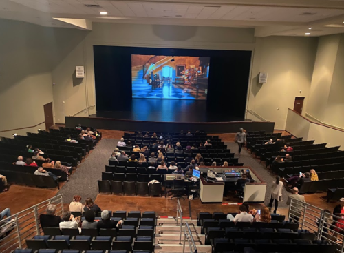 A nearly filled auditorium with an empty stage and a large screen displaying an image, audience seated and waiting.