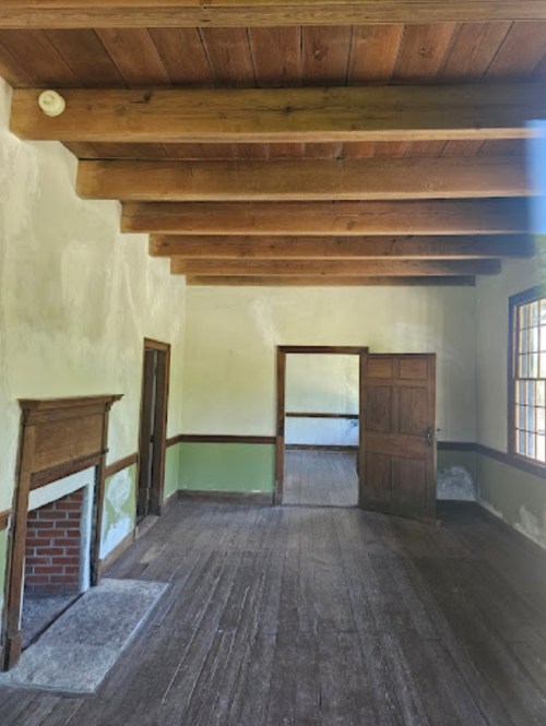 Interior of a rustic room with wooden beams, a fireplace, and two doorways, featuring peeling green walls and wooden floors.