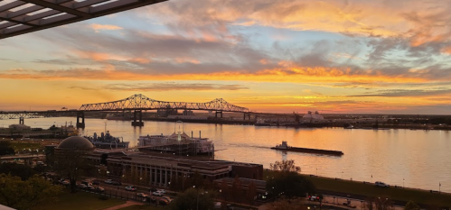 A vibrant sunset over a river, with a bridge in the background and boats on the water, reflecting colorful skies.
