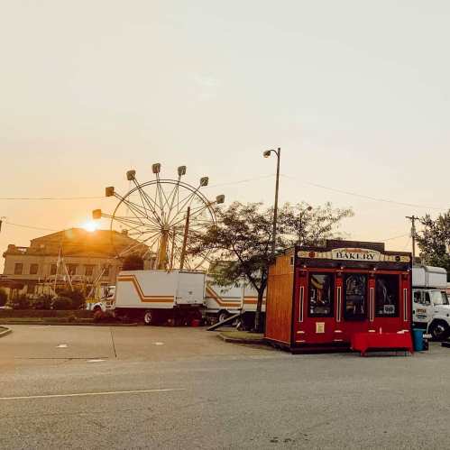 A red bakery stand in a parking lot with a ferris wheel and sunset in the background.