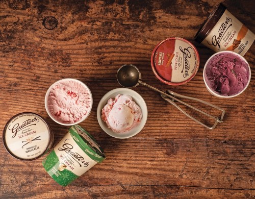 Assorted tubs of Graeter's ice cream on a wooden table, with a scoop and a bowl of strawberry ice cream.