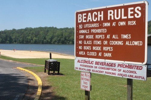 Sign displaying beach rules, including no lifeguards, no animals, and alcohol prohibition, near a calm lake.