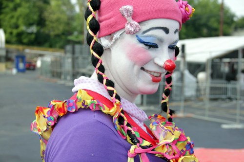 A smiling clown with colorful hair and makeup, wearing a purple outfit adorned with bright, ruffled decorations.