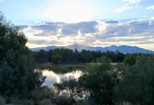 A serene landscape featuring a reflective lake surrounded by trees, with mountains and clouds illuminated by sunlight.