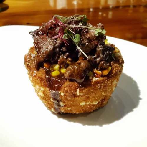 A crispy, golden-brown dish topped with savory meat, corn, and microgreens, served on a white plate.