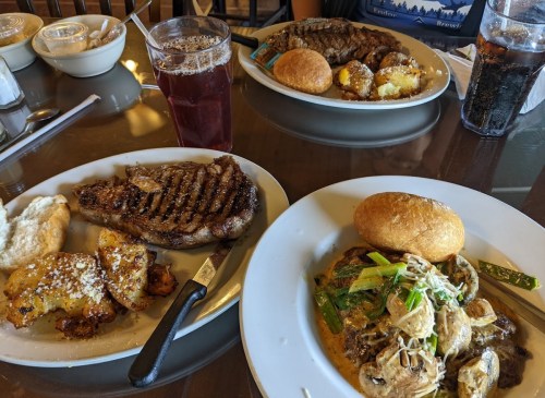 A table with two plates of food: one with steak and potatoes, the other with pasta and a roll, plus a drink.