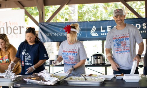 Three people serving food at an outdoor event, wearing casual shirts and gloves, with a banner in the background.