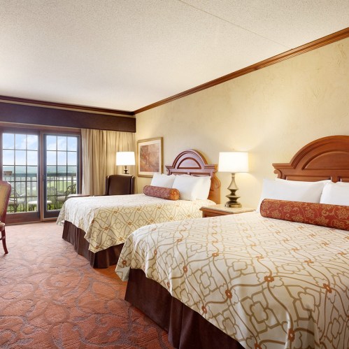 A hotel room featuring two beds, a chair, and large windows with a view, decorated in warm tones.