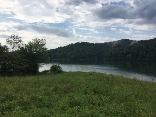 A serene lake surrounded by lush greenery and hills under a cloudy sky.
