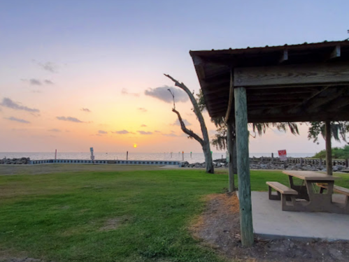 A serene beach scene at sunset, featuring a picnic area under a shelter and a calm water view.