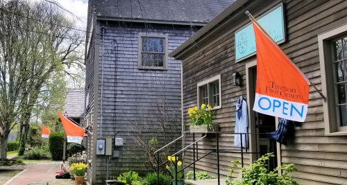 Two orange "OPEN" flags outside a wooden building, with yellow flowers in pots and a person sitting nearby.