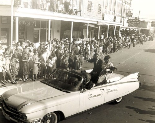A vintage parade scene with a crowd watching as a convertible car passes by, featuring a couple waving from inside.