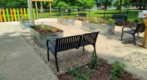 A park area with raised garden beds filled with flowers, a bench, and gravel pathways surrounded by greenery.