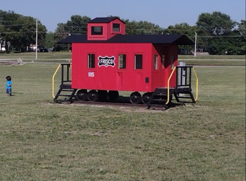 A red train caboose with "Frisco" logo sits on grass, with a child playing nearby in a park setting.
