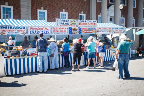 A busy food market with people browsing various food stalls, including crab cakes and Greek salads, under a sunny sky.