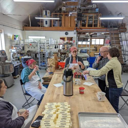A group of people in a kitchen setting, gathered around a table with coffee and baking supplies, engaged in conversation.