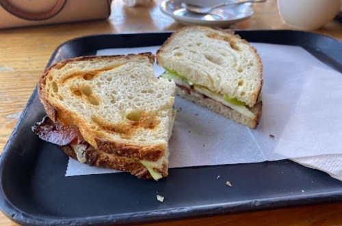 A sandwich on a black tray, cut in half, with layers of meat, cheese, and green apple slices on toasted bread.