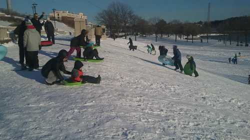 A snowy hill filled with people sledding, some sitting on sleds while others prepare to go down. Trees and buildings in the background.